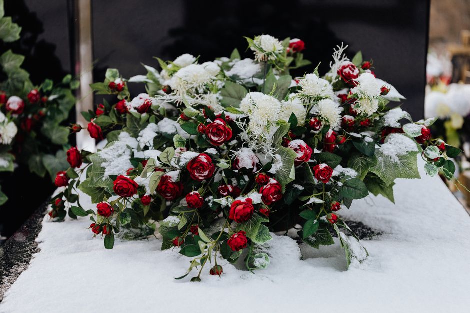 A floral arrangement featuring a mix of red roses and white chrysanthemums, accented with green holly leaves. The bouquet is dusted with a light layer of snow, highlighting the freshness of the petals