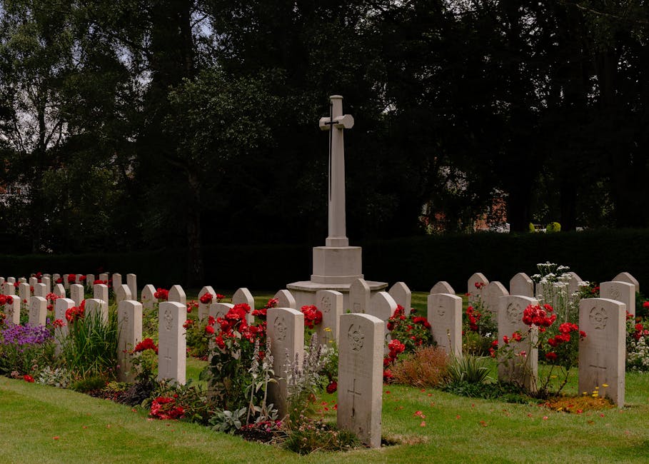 A graveyard scene featuring neatly arranged white tombstones, each engraved with symbols and inscriptions, surrounded by a vibrant array of fresh flowers including red roses, purple lavender, and whit