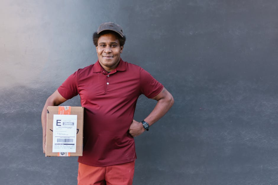 A delivery driver from flowerdeliverycranham.co.uk posing in front of a plain, neutral-colored wall while holding a small cardboard parcel with a printed shipping label. The driver, a man with dark sk